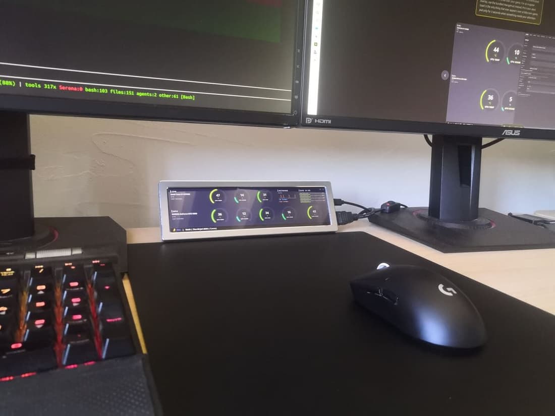 A small stretched Thermal Canary panel sitting on a real desk in front of dual monitors, next to a mechanical keyboard and gaming mouse